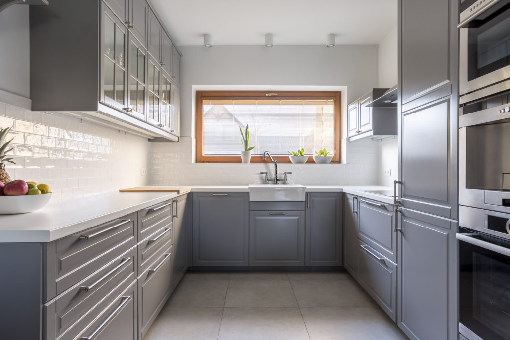 This kitchen design shows light grey cabinetry, a white countertop and a white farmhouse sink under a large window. There’s a bowl of fruit on the counter, and there’s white subway tile used as the backsplash in the space.