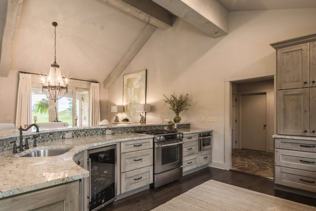 This large kitchen design features distressed, white cabinetry with a white and grey marble countertop. There’s a tiled backsplash, a white rug, a wine cooler and a vase featured in the space, too.