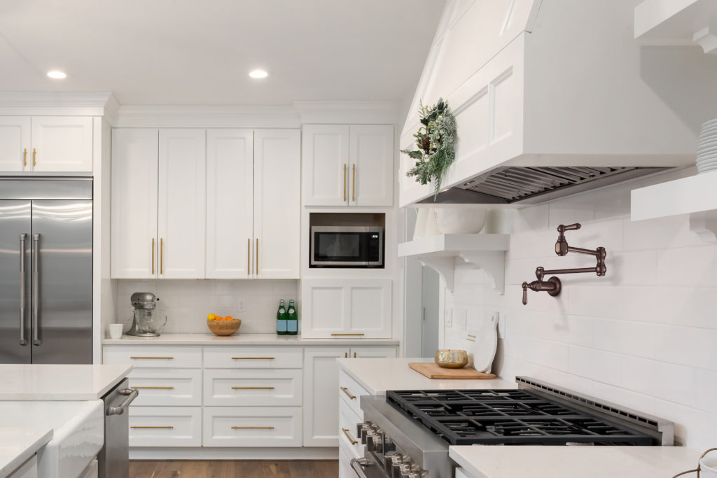 This all white kitchen design has white cabinetry, white countertops and white backsplashes throughout the design. There’s gold hardware in the space, and there’s a bronze pot filler located above the stove.