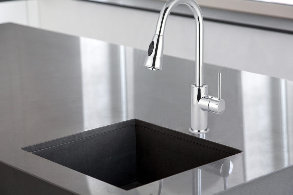 This chrome faucet has a single handle design. The square sink is within the dark grey island.