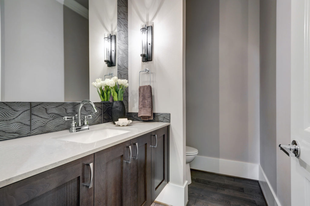 This bathroom design features a large vanity with a sink. The cabinetry is a dark wood while the backsplash is a dark grey and white patterned. There is a brown towel hanging on the towel bar and a black vase holding white stemmed flowers in the corner of the vanity.