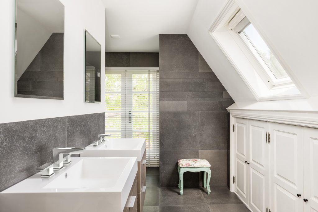 This small bathroom has white sinks and white cabinetry. There is a dark, tiled wall in the space and a light green stool sitting against the wall.