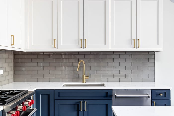 Image of a white, brass, and navy blue kitchen. The faucet is single-handed brushed brass and matches the cupboard hardware. The top cupboards are white and the bottom cupboards are navy. The counter is white granite and the stove and dishwasher are silver.