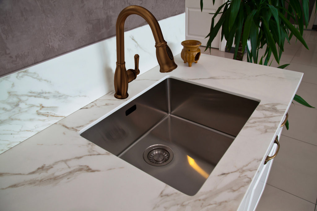 This sink design has a copper faucet and a silver sink. There’s a marble countertop and a grey backsplash.