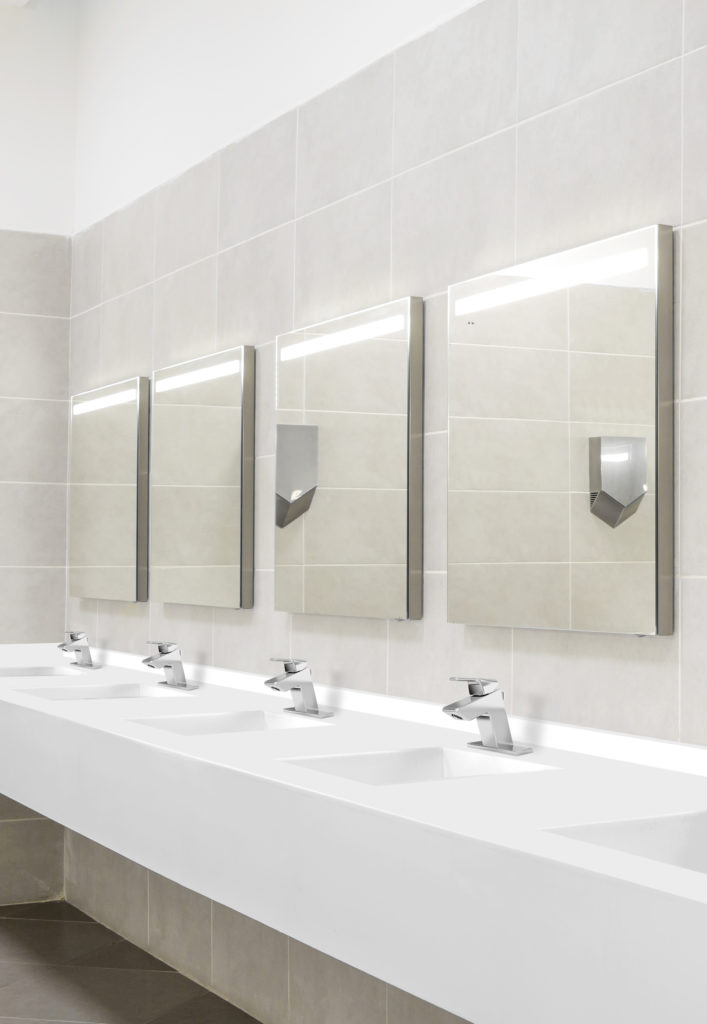 This bathroom has multiple sinks with silver faucets. The countertop and sinks are white, and there are tan tiles on the flooring and the walls.