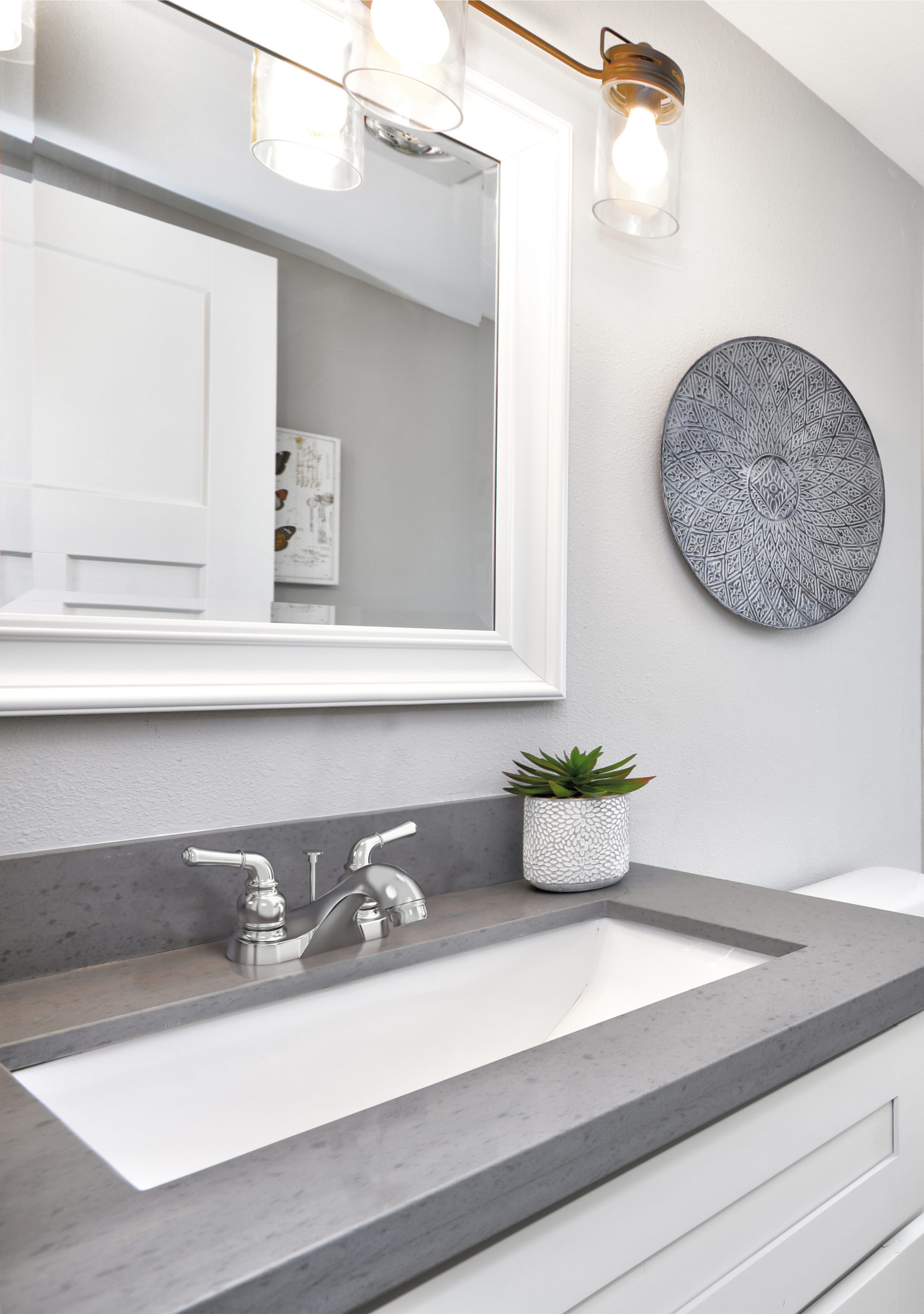 This vanity features white cabinetry, a grey countertop, silver faucet, a white-framed mirror, a plant and wall art.