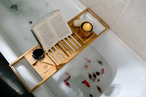 This bathroom design has a large, white tub filled with bubbles and rose petals. The tub also has an expandable bathtub caddy with a glass of wine, a candle and a book placed on it.
